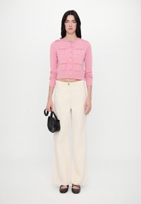 Light pink cropped cardigan with button details and two patch pockets, paired with cream wide-leg trousers and a small black handbag.