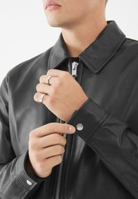 Hombre con chaqueta de cuero negra ajustando el puño abotonado, mostrando dos anillos de plata en los dedos, primer plano del torso y las manos.