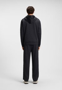 Felpa nera in pile con cappuccio, indossata con pantaloni sportivi neri abbinati. Entrambi presentano un motivo texturizzato e polsini elastici alle caviglie e ai polsi.