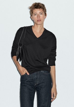 Maglione nero lavorato a maglia con scollo a V e maniche lunghe, abbinato a jeans in denim scuro. La modella tiene una borsa nera con texture.