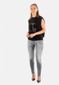 Top negro de punto con un logo brillante, combinado con vaqueros ajustados de color azul claro y tacones altos negros, llevado por una modelo de pie.