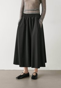 Gonna nera alla caviglia con vita elasticizzata arricciata; tessuto liscio; due tasche laterali; abbinata a scarpe nere basse.