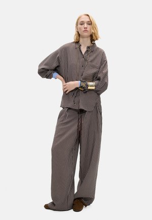 Osoba ubrana w luźną, brązową, pasiastą koszulę zapinaną na guziki i pasujące szerokie spodnie z brązowymi butami, stojąca z jedną ręką na biodrze.