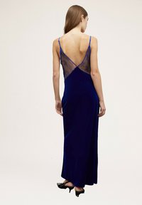 Vestido de noche de terciopelo en azul profundo con espalda en forma de V, adornado con detalles de encaje transparente. Presenta tirantes finos y una falda larga hasta el suelo.