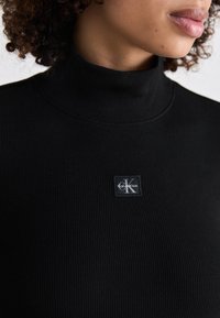 Black ribbed turtleneck sweater made of stretchy material, featuring a square logo patch on the chest with a white "K" and brand name.