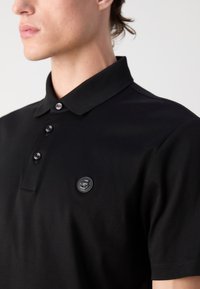 Black polo shirt made of soft cotton. Features a classic collar, three buttons, and a textured logo patch on the left chest.