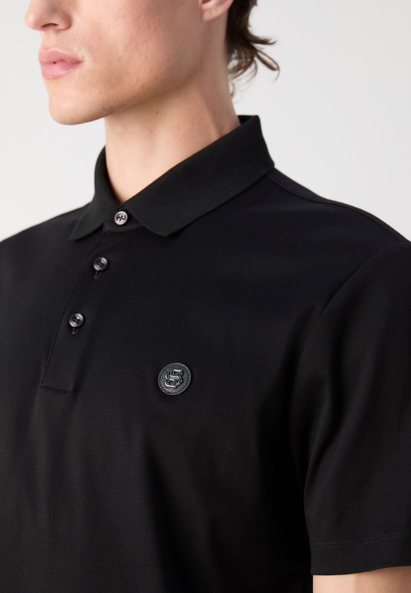 Schwarzes Polo-Shirt aus weichem Baumwollstoff. Verfügt über einen klassischen Kragen, drei Knöpfe und ein strukturiertes Logo-Patch auf der linken Brust.