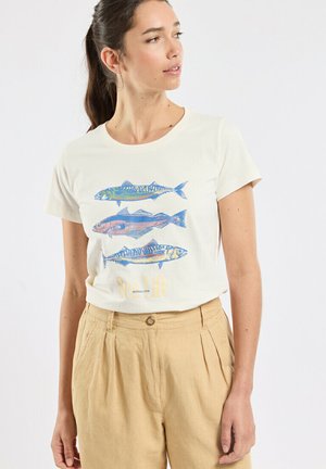 Vrouw draagt een wit T-shirt met drie kleurrijke visillustraties en beige broek met hoge taille, kijkt naar links.