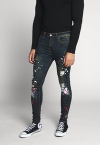 Jeans skinny de denim con diseño de salpicaduras de pintura, que presentan áreas desgastadas, color azul oscuro y acentos de pintura roja y blanca.