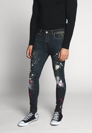 Denim skinny jeans met een spetterverfontwerp, featuring gescheurde delen, donkerblauwe kleur en contrasterende rode en witte verfaccenten.