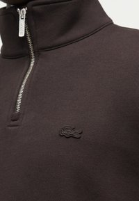 Σκούρο καφέ πουλόβερ με φερμουάρ στο ένα τέταρτο και ασημένιο φερμουάρ, με μικρό κεντημένο λογότυπο κροκόδειλου στο αριστερό μέρος του στήθους.