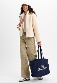 Fleece jacket in roomkleur met een ritssluiting en twee voorzakken, gecombineerd met olijfgroene cargobroek en een marineblauwe tote bag met witte tekst. Witte sneakers.