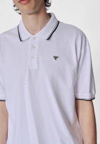 Witte polo met een zwarte gestreepte kraag, drie knopen en een klein geborduurd bijenlogo op de borst. De stof lijkt textuur te hebben.