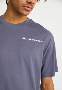 Pelēka kokvilnas T-krekls ar apaļu kakla izgriezumu, īsām piedurknēm un BMW Motorsport logo ar ziliem un sarkaniem akcentiem uz krūškurvja.