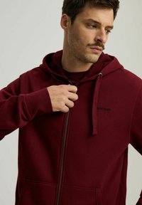 Bordo bluza z kapturem na zamek, wykonana z miękkiego materiału, z kieszenią kangurką i regulowanym kapturem. Czarna logo na klatce piersiowej. Gładka tekstura, casualowy design.