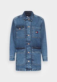 Denim jacket in medium blue with a button-up front, four pockets, contrasting stitching, and a small logo patch on the chest pocket.