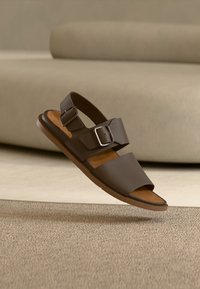 Καφέ δερμάτινο σανδάλι με δύο ρυθμιζόμενους ιμάντες και ασημένιες αγκράφες. Μαλακό ταμπά πέλμα και ευέλικτη λαστιχένια σόλα. Μινιμαλιστικός σχεδιασμός.