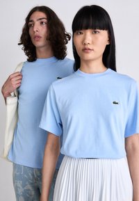 Zwei Personen tragen hellblaue Lacoste-T-Shirts, eine mit einem weißen, plissierten Rock, die andere hält eine cremefarbene Tragetasche, beide stehen vor einem schlichten Hintergrund.
