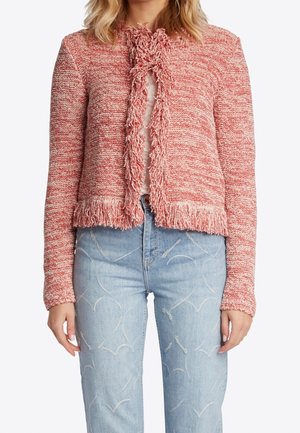 Mujer con chaqueta roja texturizada con flecos sobre una camiseta blanca y vaqueros azul claro con patrones cosidos.