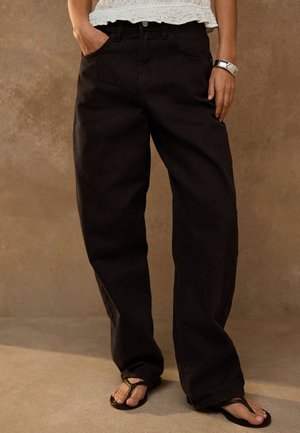 Personne portant un pantalon noir ample, un haut blanc texturé, des sandales noires à bride, une main dans la poche et un bracelet argenté au poignet.