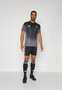 Maglia atletica grigia con pantaloncini neri, caratterizzata da accenti gialli, motivi diagonali e logo. Abbinata a calzini neri e scarpe con tacchetti.