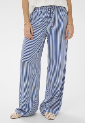 Voľné modro-biele nohavice s vertikálnymi pruhmi a šnúrkou v páse, nosené s bielymi pletenými slip-on topánkami a zlatými náramkami na rukách.