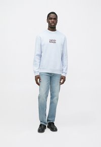 Világoskék pulóver kerek nyakkivágással, barna "Calvin Klein" logóval. Világoskék farmerrel és fekete cipővel viselve.