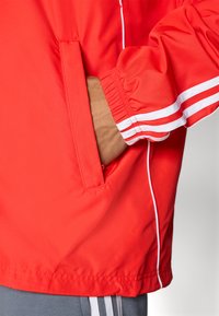 Osoba v červenej bunde s bielymi pruhmi na manžetách a bočných liniek, ruka vložená do predného vrecka, spodná časť sivých nohavíc viditeľná.