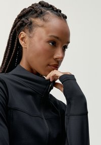 Czarna sportowa kurtka na zamek z wysokim kołnierzem, z detalami szycia i gładką teksturą. Zamek z przodu umożliwia regulowane rozpięcie.