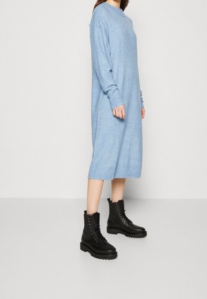 Femme portant une robe longue en maille bleu clair associée à des bottes de combat noires à lacets, posant devant un fond gris clair uni.