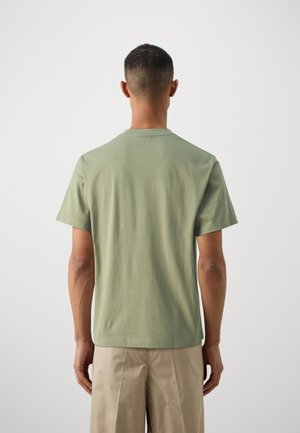 Ein Mann steht mit dem Rücken zur Kamera, trägt ein schlichtes hellgrünes T-Shirt mit kurzen Ärmeln und beigen Hosen vor einem neutralen Hintergrund.