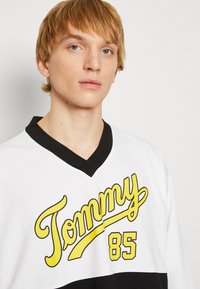 Maglietta bianca con accenti neri, che presenta il testo giallo "Tommy" e il numero "85". Design a V, tessuto testurizzato.