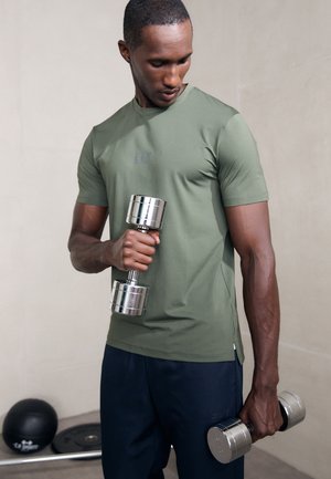 Green athletic shirt with short sleeves, holding silver dumbbells. The background features weights and a fitness mat, emphasizing a workout setting.