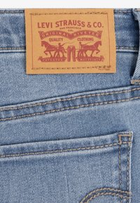 Δερμάτινη ετικέτα της Levi Strauss & Co. σε τζιν, σε καφέ χρώμα με κόκκινο ανάγλυφο κείμενο και απεικονίσεις αλόγων και μορφών, τζιν με ανοιχτό μπλε απόχρωση.