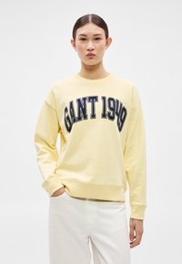 Sárga pulóver kerek nyakkivágással és ejtett vállakkal, elöl vastag tengerészkék felirattal: "GANT 1949". Simább anyag textúra.