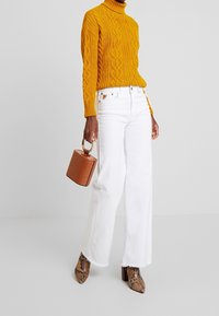 Gelbes grobstrickiges Rollkragensweatshirt, weiße weit geschnittene Jeans, braune Handtasche mit Krokodilprägung und gemusterte Absatzstiefel.