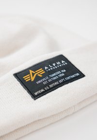 Kremasti pleteni zimski šešir s crnom pravokutnom etiketom koja sadrži žute i bijele natpise i logo. Etiketa označava marku i detalje o proizvodnji.