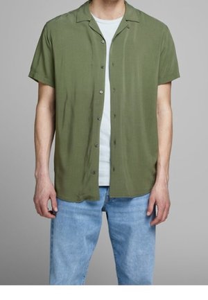 Homme portant une chemise à manches courtes vert olive déboutonnée sur un t-shirt blanc avec un jean bleu clair, sur un fond gris.