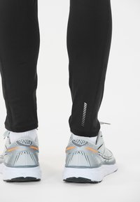Zwarte sportleggings met een reflecterende lijn, gecombineerd met lichtgrijze hardloopschoenen met oranje "ENDURANCE" accenten en een mesh textuur.