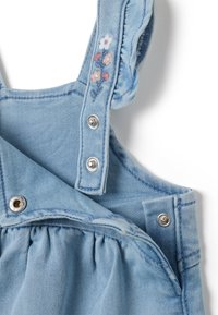 Pantalón de mezclilla azul claro con acentos florales bordados en las correas de los hombros; cuenta con broches de presión plateados y detalle de cintura fruncida.