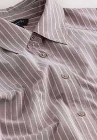 Chemise marron clair à fines rayures verticales roses, boutonnière devant et col pointu, posée à plat sur une surface blanche.