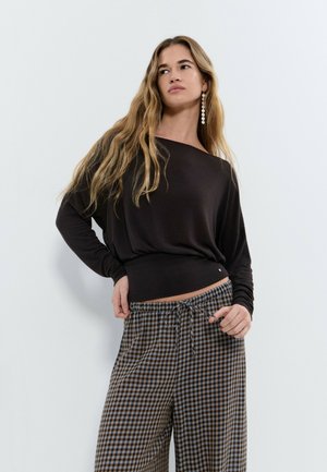 Femme aux longs cheveux blonds portant un haut noir à manches longues et un pantalon large à carreaux marron et bleu sur un fond clair uni.