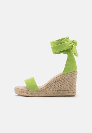 Grüne Keilsandale aus Wildleder mit Knöchelwickelriemen und geflochtener Jute-Plateausohle vor weißem Hintergrund.
