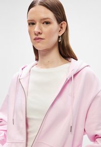 Svetloružová mikina na zips s kapucňou a šnúrkou, pod ktorou je jemný biely pružný golier. Diskrétny logo detail v zodpovedajúcej farbe.