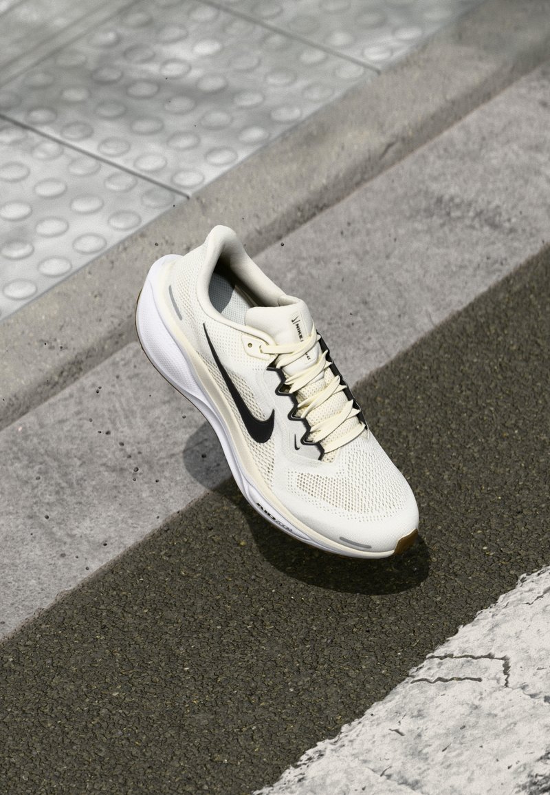 Nike running shoe in off-white mesh with a black swoosh. Features a textured sole and light-colored laces, positioned on a textured surface.