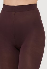 Tmavě vínové legíny s hladkou, pružnou texturou a širokým žebrovaným pasem; mají bezešvou konstrukci v oblasti vnitřní strany stehen.