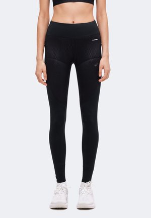 Donna con leggings neri strutturati e scarpe da ginnastica bianche, in piedi con le braccia rilassate lungo i fianchi davanti a uno sfondo uniforme.