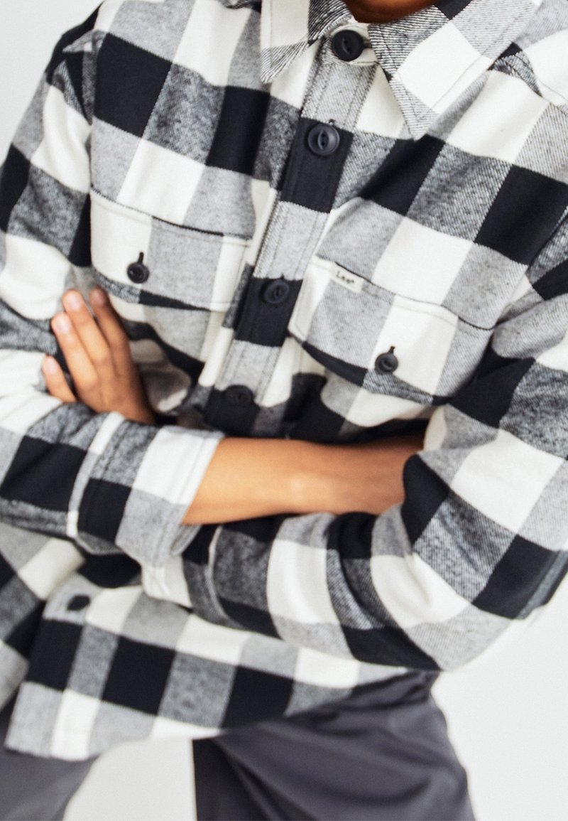 Black and white checkered flannel shirt with a collar, featuring large buttons, two chest pockets, and contrasting patterns on sleeves.
