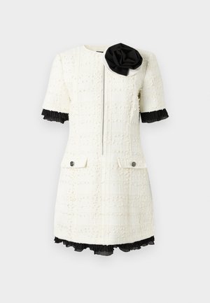 Robe blanche texturée à manches courtes avec fermeture éclair à l'avant, bordure plissée noire sur les manches et l'ourlet, deux poches avant, et grande fleur en tissu noir et blanc sur l'épaule.
