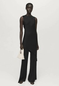 Zwarte mouwloze top met een asymmetrische zoom, gecombineerd met brede zwarte broek. Witte handtas in de hand en zwarte sandalen aan de voeten.